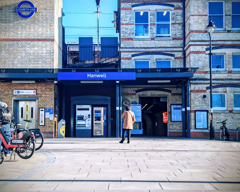 A person in a beige jacket and dark trousers stands outside the entrance of Hanwell station, part of the Elizabeth Line, situated in a brick building with large glass windows. The station entrance features a dark canopy with a blue sign displaying the station name. To the left of the entrance, there are electronic ticket machines and a bike rack with actual bikes parked, including those with Uber and Vaux logos. On the right side, there are information boards mounted on the brick wall, along with a black lamppost and a bicycle secured to a bike rack. The pavement is paved with square tiles, and the area is well-lit with natural daylight. This scene depicts a typical urban setting where a professional moving service, like Man and Van Tottenham Hale, might load furniture or packing boxes into a van for home relocation or furniture transport, as part of their removals process.