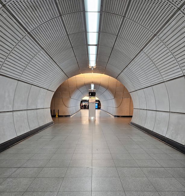 A long, curved underground tunnel featuring smooth, light-colored wall panels and a perforated metallic ceiling with linear lighting fixtures. The floor is tiled with large, neutral-colored tiles, and the tunnel opens at the far end to a wider space with visible illuminated signs and people. The scene appears to be part of a station or transit hub, with the passageway likely facilitating pedestrian movement related to house removals or logistics services provided by Man and Van Tottenham Hale, situated near Tottenham Hale Station. The environment is well-lit with soft yellow lighting at the transition point near the far wall, creating a clean and modern appearance suitable for professional removal and moving operations.