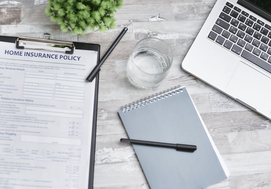 A woman with long black hair, wearing a grey checkered vest over a white shirt, is holding a clipboard with a home insurance policy form and a pen, while speaking to a man with short brown hair, dress