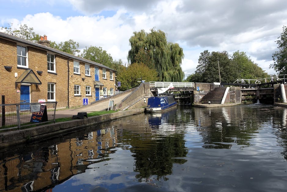 A canal scene with a brick residential building on the left, featuring white-framed windows and a blue door, next to a small fenced garden area. There is a metal footbridge crossing over the lock, with steps leading down on both sides, and a narrowboat moored alongside the canal wall. Several cardboard boxes and bundled packages wrapped in plastic and fabric are placed on the pavement near the building, suggesting packing and home relocation activities. The scene is set outdoors during daytime with a partly cloudy sky, and a large green tree in the background. The environment indicates a residential area involved in a furniture transport or moving process, with visible elements suitable for house removals services by Man and Van Tottenham Hale, emphasizing loading and packing logistics in an urban setting.
