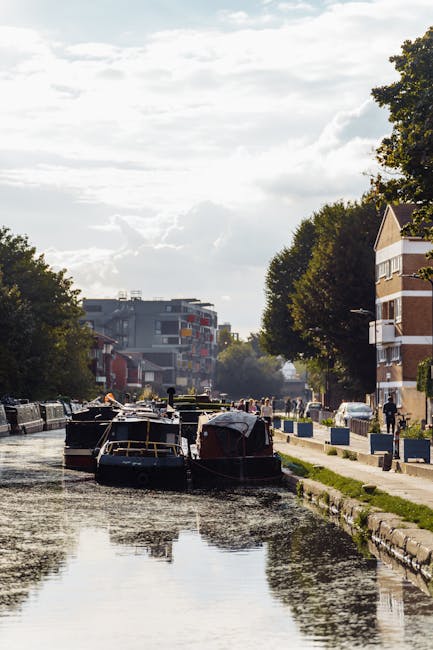 The image shows a canal scene with several narrowboats moored along the water's edge, some covered with tarpaulins and others visible with wooden and metal structures. The canal is lined with a paved walkway and greenery, including trees with dense foliage on both sides. In the background, there are residential buildings and modern apartment complexes, with some multi-storey structures featuring balconies. The sky is partly cloudy with sunlight filtering through, casting reflections on the canal surface. The scene is calm and typical of urban house relocation or moving logistics in the Tottenham Hale area, with the canal possibly used to assist in transporting furniture or belongings during a home removal process. The overall environment suggests a residential neighbourhood with infrastructure suitable for loading and unloading during a house move, as handled by companies like Man and Van Tottenham Hale.