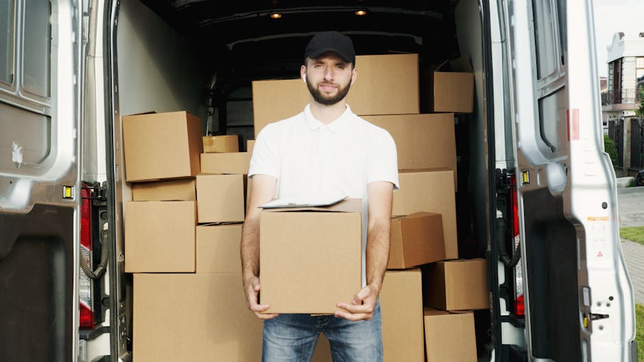 A man wearing a black cap, white t-shirt, and blue jeans is seated on the pavement beside the open rear door of a white van, which is filled with stacked cardboard boxes of various sizes used for home